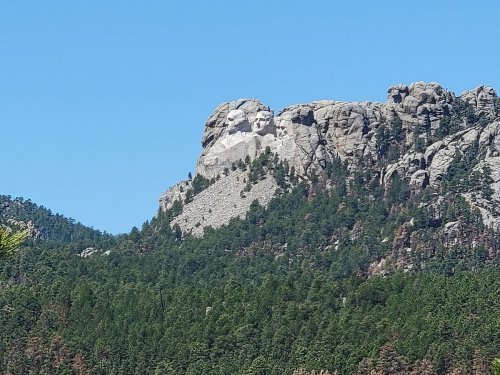 A mountain landscape featuring a large rock formation resembling a face, surrounded by dense green trees and a clear blue sky.
