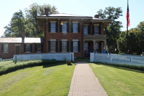 A historic brick house with a white picket fence, surrounded by green grass and trees, under a clear blue sky.