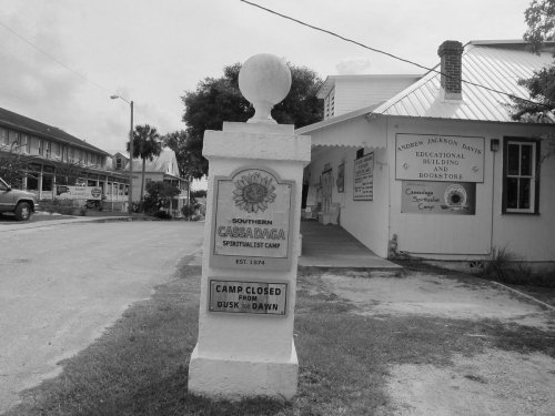 Sign for Southern Cassadaga Spiritualist Camp, established in 1874, with a note about camp hours. Black and white image.
