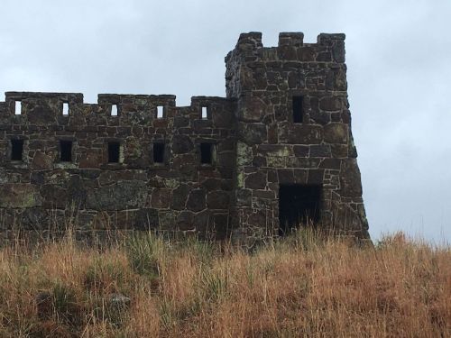 A stone tower with small windows, set against a cloudy sky and surrounded by tall grass.