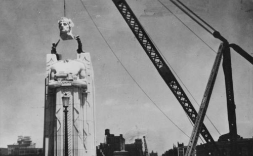 A historic black-and-white photo of a statue's head being lifted by a crane, with a city skyline in the background.