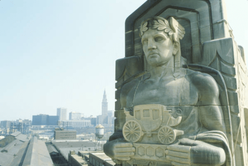 A large stone statue of a man holding a carriage, with a city skyline in the background.