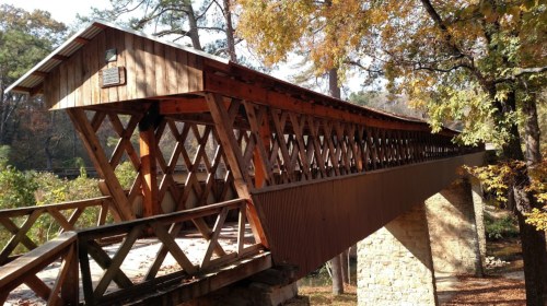 A wooden covered bridge with a lattice design, surrounded by trees and autumn foliage.