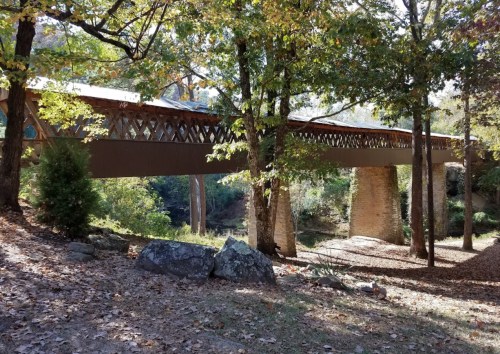 A wooden bridge spans a river, surrounded by trees with autumn foliage and rocky terrain.