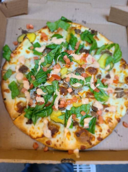 A pizza topped with lettuce, pickles, tomatoes, and a creamy sauce, served in a pizza box.
