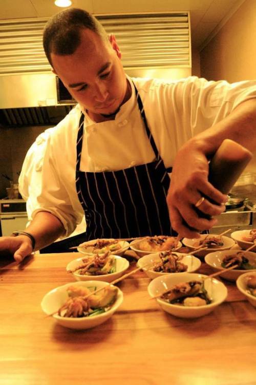 A chef in a striped apron carefully plates food in small bowls in a kitchen setting.