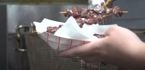 A hand holds a paper tray filled with skewered meat, ready to be served from a fryer.