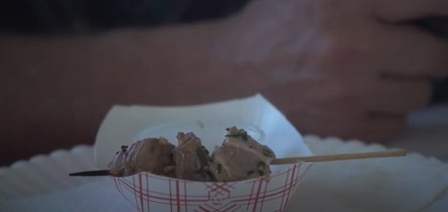 A close-up of a small serving of skewered meat on a paper tray, with a hand holding a phone in the background.