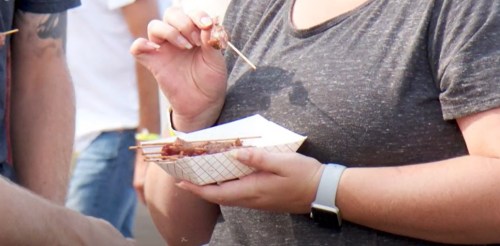 A person holds a plate of skewered food, taking a bite while others are seen in the background.