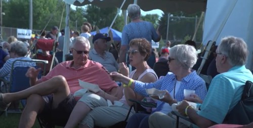 A group of people enjoying food and conversation at an outdoor event under a tent, with others in the background.