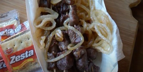A serving of grilled meat skewers with sautéed onions, accompanied by packs of Zesta saltine crackers.