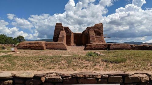 Ancient adobe ruins under a blue sky with fluffy clouds, surrounded by grass and distant hills.