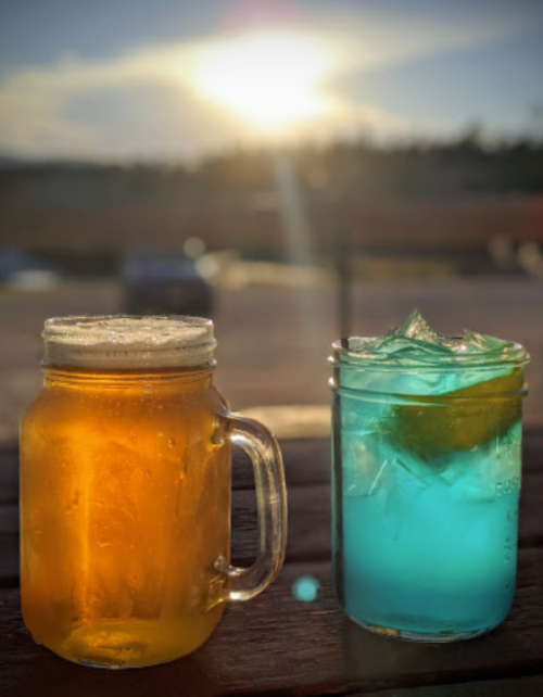 Two drinks sit on a table: a frothy amber beer in a jar and a blue cocktail with ice and a lime slice, with a sunset in the background.