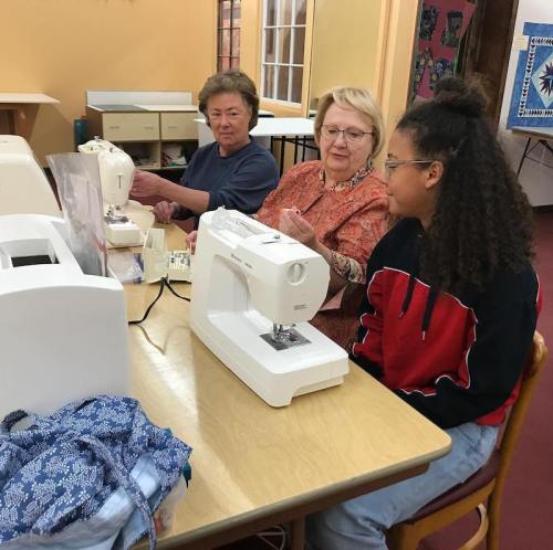 Three people sit at a table with sewing machines, engaged in a sewing activity in a bright, cozy room.