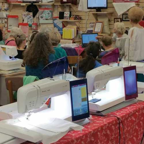 A sewing class in a bright room with participants using sewing machines and computers, surrounded by fabric and supplies.
