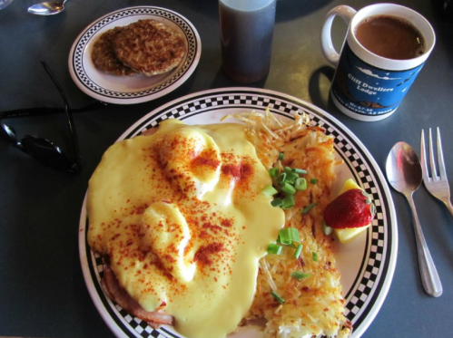 A plate of eggs Benedict with hash browns, a side of pancakes, coffee, and sunglasses on a table.