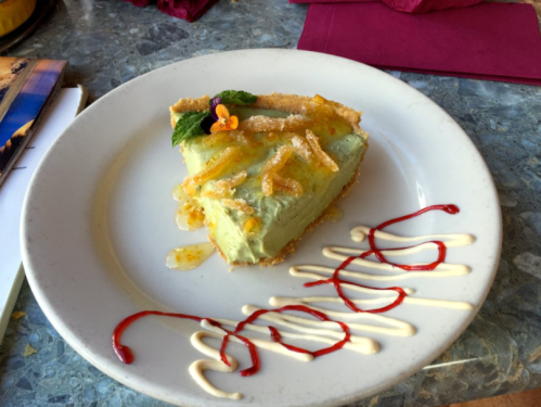 A slice of green dessert pie topped with orange zest and a flower, drizzled with red and white sauces on a plate.