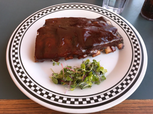 A plate of barbecue ribs covered in sauce, served with a side of microgreens on a checkered rim plate.
