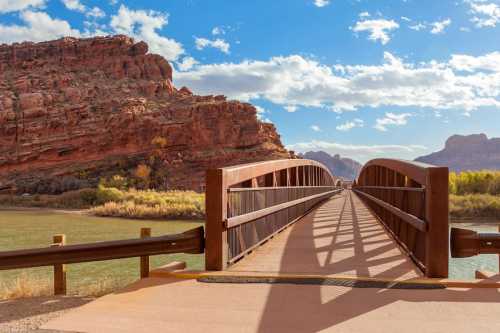 A wooden bridge spans a river, surrounded by red rock formations and a blue sky with scattered clouds.