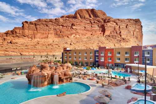 A resort with pools and waterfalls, set against a backdrop of a large red rock formation under a blue sky.