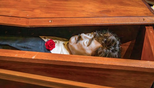 A lifelike figure of a man in a coffin, wearing a suit and a red rose on his lapel, inside a wooden casket.