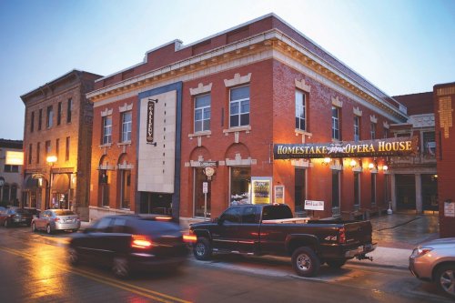Historic brick building with "Homestake Opera House" sign, streetlights illuminating the evening scene. Cars parked outside.