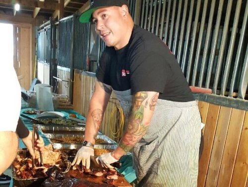 A man with tattoos prepares food at a barbecue, smiling while serving from a large tray in a rustic setting.