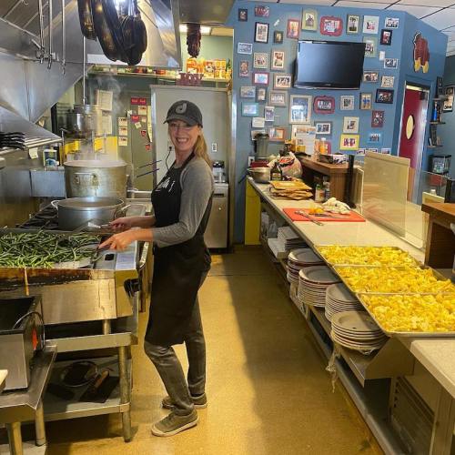 A smiling chef in an apron prepares food in a busy kitchen, with dishes and colorful decor in the background.