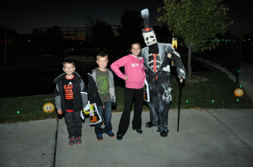Three children pose with a spooky costumed figure at dusk, surrounded by Halloween decorations.
