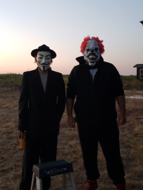 Two people in costumes: one wears a Guy Fawkes mask and suit, the other a clown mask with red hair, standing outdoors at sunset.