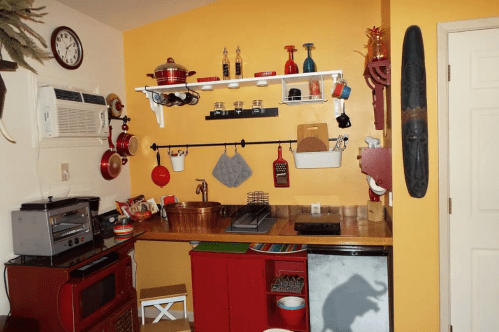 A cozy kitchen with yellow walls, red accents, and various cooking utensils displayed on shelves.