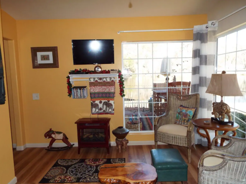 Cozy living room with yellow walls, a TV, decorative shelves, and a rocking horse, featuring large windows and wooden furniture.