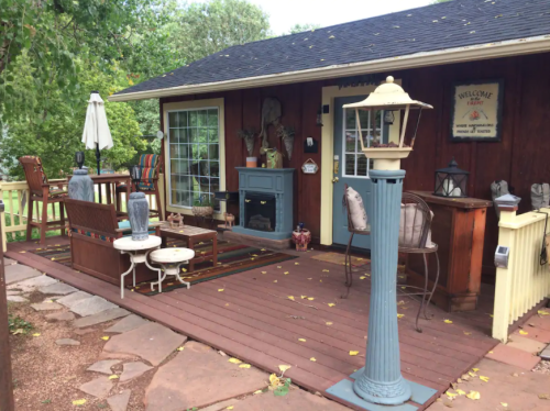 A cozy outdoor patio with seating, a lantern, and decorative elements, surrounded by greenery and a wooden cabin.