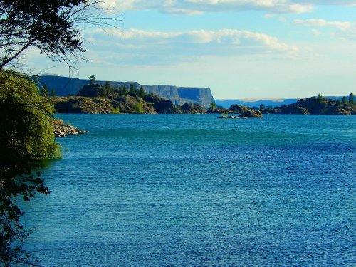 A serene lake surrounded by rocky cliffs and lush greenery under a blue sky with scattered clouds.