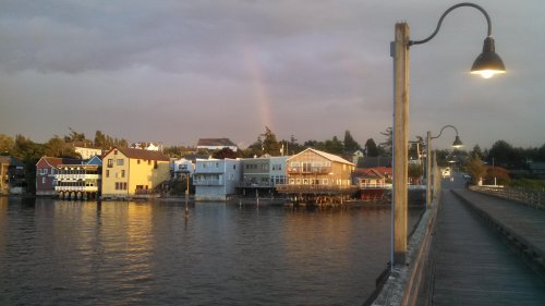 A scenic waterfront view with colorful houses, a rainbow in the sky, and a wooden walkway along the water.