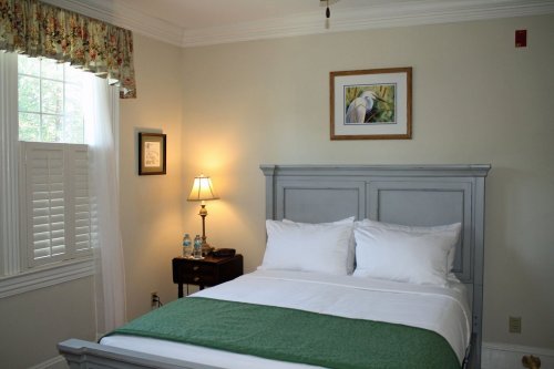Cozy bedroom with a gray bed, white linens, a lamp, and a framed artwork on the wall. Natural light from the window.