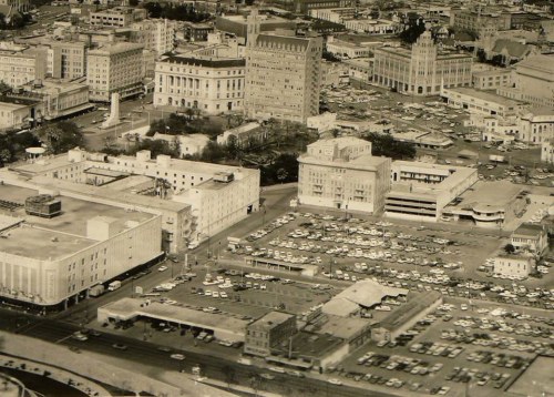 Aerial view of a cityscape featuring buildings, parking lots, and streets, captured in black and white.