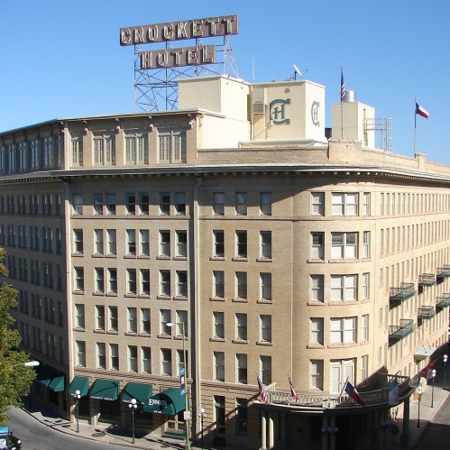 Historic Crockett Hotel building with a rooftop sign, featuring multiple floors and green awnings. Clear blue sky above.