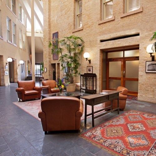 A spacious lobby with leather chairs, a wooden table, plants, and decorative rugs, featuring large windows and warm lighting.