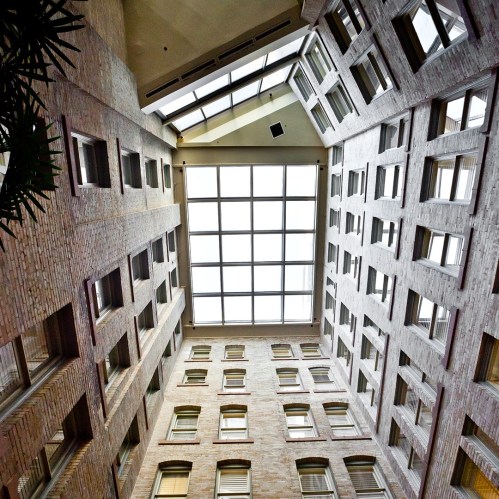 Looking up at a glass ceiling surrounded by brick walls and windows in a spacious atrium.