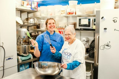 A young woman and an elderly woman smile in a kitchen, holding a whisk and spatula, ready to cook together.