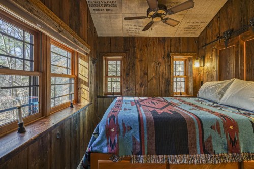 Cozy wooden bedroom with a patterned blanket, windows letting in natural light, and a ceiling fan.