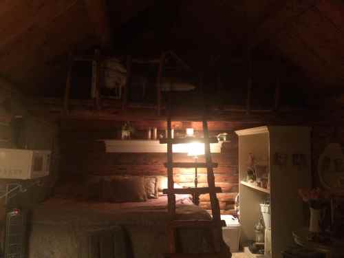 Cozy cabin interior with a loft, wooden ladder, and warm lighting, featuring a bed and rustic decor.