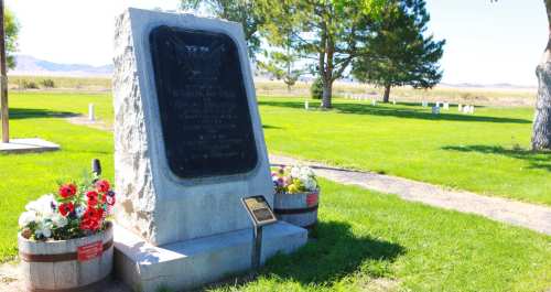 A stone monument with a plaque, surrounded by flowers, in a grassy area with trees and distant gravestones.