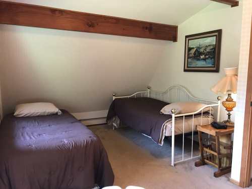 A cozy bedroom with two twin beds, a wooden beam, a lamp, and a framed painting on the wall.