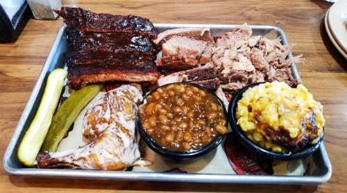 A BBQ platter featuring ribs, pulled pork, smoked chicken, baked beans, mac and cheese, and pickles on a tray.