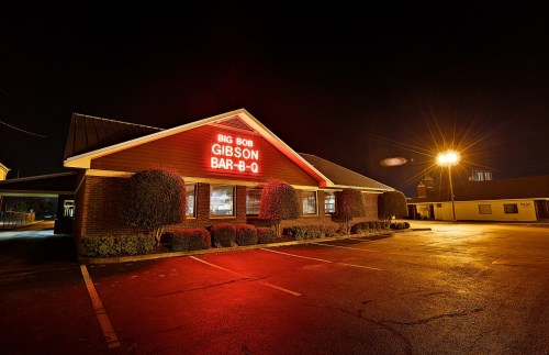A barbecue restaurant at night, featuring a lit sign that reads "Big Bob Gibson Bar-B-Q" and well-trimmed bushes.