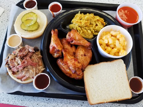 A tray with BBQ chicken wings, pulled pork sandwich, macaroni and cheese, pickles, and sides of sauce.