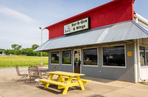 Exterior of a barbecue and wings restaurant with a red roof, large windows, and a yellow picnic table outside.
