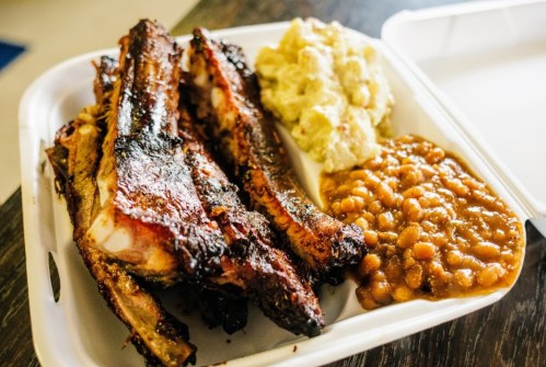 A takeout container with grilled ribs, potato salad, and baked beans.